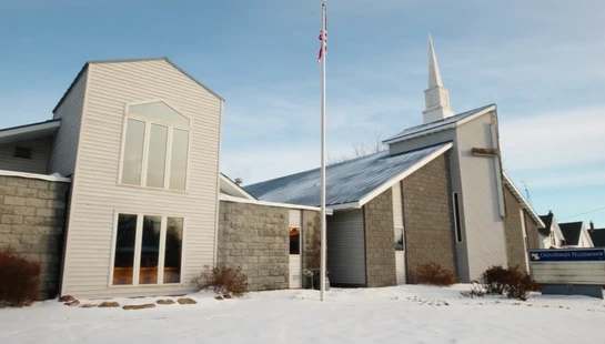 A modern church with white siding and stone accents sits in a snowy landscape under a blue sky. A sign reads "Crossroads Fellowship" and a US flag flies.